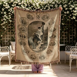 May include: A beige woven throw blanket featuring a large central illustration of the crucifixion surrounded by twelve smaller circular vignettes depicting scenes from the Stations of the Cross, finished with a decorative fringed border.