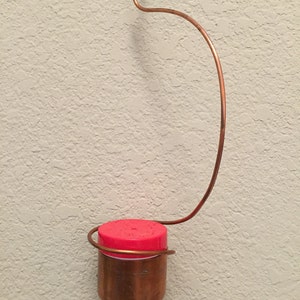 May include: A copper hummingbird feeder with a red plastic top hanging from a copper wire hook.
