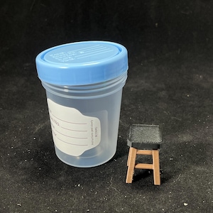 May include: A clear plastic specimen container with a light blue lid and a white label. Beside it is a miniature brown wooden stool with a black textured seat. The background is black.