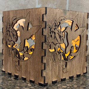May include: A wooden candle holder with a cutout design of hummingbirds and flowers. The candle holder is lit from within, casting a warm glow.