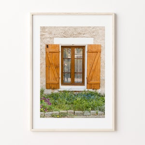 Puede incluir: Una ventana con contraventanas de madera en un edificio de piedra. Las contraventanas están cerradas y pintadas de un color marrón cálido. Hay plantas verdes y flores creciendo frente a la ventana.