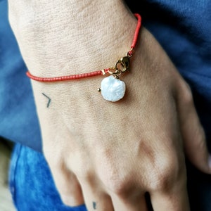 Könnte beinhalten: Ein rotes Perlenarmband mit einem goldenen Verschluss und einem weißen Perlenanhänger.