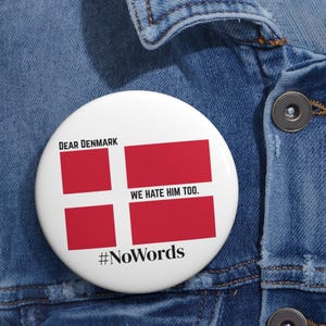 Op de afbeelding: Een witte button met het Deense vlagontwerp, een rood kruis op een witte achtergrond. Tekst bevat "DEAR DENMARK", "WE HATE HIM TOO." en "#NoWords". De button is vastgemaakt aan een spijkerjasje.