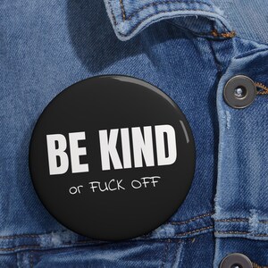 Può includere: Un bottone nero con testo bianco che recita "BE KIND" e "or FUCK OFF" in uno stile di scrittura a mano.