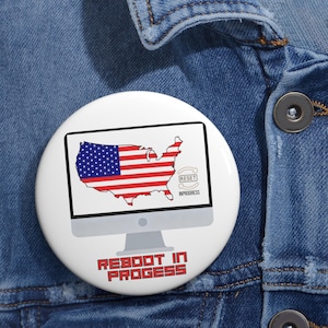May include: A white button badge with a computer screen design showing the U.S. map in the American flag colours. The screen displays "RESET IN PROGRESS". The badge also has the words "REBOOT IN PROGRESS" in red.