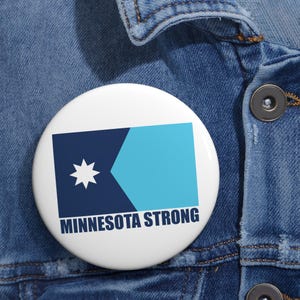 May include: A white button pin with a design featuring a dark blue and light blue geometric shape and a white star. The text "MINNESOTA STRONG" is printed in dark blue below the design. The pin is set against a denim background.