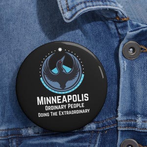 Pode incluir: Um botão redondo preto com o texto "MINNEAPOLIS ORDINARY PEOPLE DOING THE EXTRAORDINARY". O botão apresenta um design estilizado de pássaro em azul e branco, com uma estrela acima. O botão está sobre um fundo de ganga azul.