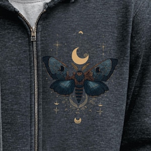 May include: Dark gray zip-up hoodie featuring a detailed illustration of a moth with blue wings and a golden body. The design includes crescent moons and stars, creating a celestial theme. The hoodie has a full zipper and a soft, textured appearance.