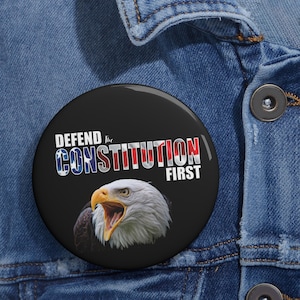Könnte beinhalten: Ein runder, schwarzer Button mit dem Text "DEFEND the CONSTITUTION FIRST" in Weiß, Rot und Blau. Der Button zeigt eine Illustration eines Weißkopfseeadlers. Der Button befindet sich vor einem Jeanshintergrund.