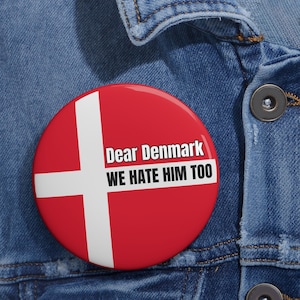 May include: A round, red button with a white cross design, resembling the Danish flag. The button has the text "Dear Denmark WE HATE HIM TOO" in white. The button is pinned to a blue denim jacket.
