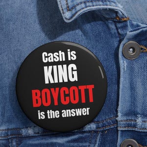 以下が含まれることがあります： 「Cash is KING BOYCOTT is the answer」という白と赤の文字が書かれた黒い丸いボタン。青いデニムジャケットにピンで留められています。