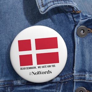 Op de afbeelding: Een witte button met de Deense vlag, een rood-wit kruis. De tekst "DEAR DENMARK, WE HATE HIM TOO." en de hashtag "#NoWords" zijn in zwart op de button gedrukt.
