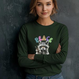 May include: Dark green long-sleeve shirt featuring a raccoon graphic giving a peace sign. The word "PEACE" is written above the raccoon in colorful bubble letters. The shirt is made of a soft material.