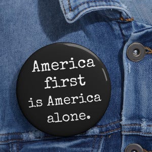 May include: A black button with white text that reads "America first is America alone." The button is round and is set against a denim jacket.
