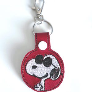 May include: Red leather keyring with a white embroidered image of Snoopy wearing sunglasses.