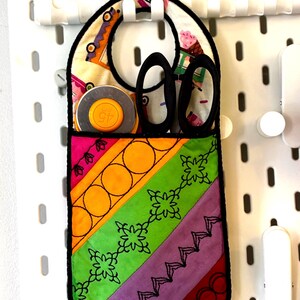 May include: A colourful fabric organiser with a black trim, hanging on a white pegboard. The organiser has a pocket for scissors, a pocket for a rotary cutter, and a pocket for other sewing supplies. The organiser is decorated with a variety of colourful fabrics and embroidery.