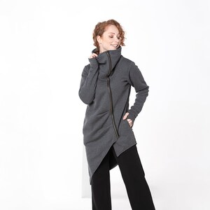 May include: A gray hooded sweatshirt with a zipper down the front and a high collar. The sweatshirt is asymmetrical and has a long, flowing design. The model is wearing black pants and yellow shoes.