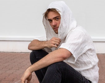 May include: A person wearing a white hooded shirt and black jeans sits on a brick surface. The shirt has short sleeves and a relaxed fit. White athletic shoes with visible air pockets complete the outfit.