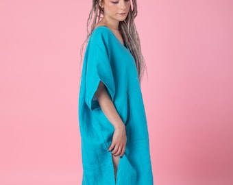 May include: A woman wearing a turquoise linen dress with a loose fit and a v-neck. She has long gray dreadlocks and is standing on a pink background.
