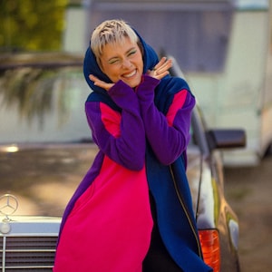 May include: A color-blocked hooded coat in blue, purple, and hot pink. The coat has a long, flowing design and is worn by a person leaning against a car. The person is smiling and wearing black shorts.