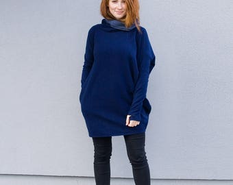 May include: A woman wearing a navy blue tunic sweater with a cowl neck and long sleeves, paired with black pants and burgundy ankle boots. The sweater has a relaxed fit and pockets.