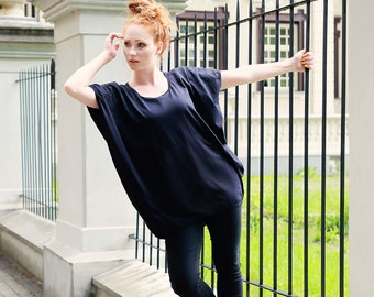 Pode incluir: Uma mulher vestindo uma blusa preta solta com decote redondo e calças pretas. Ela está em pé em frente a uma cerca de metal preta e tem o braço levantado, segurando a cerca.