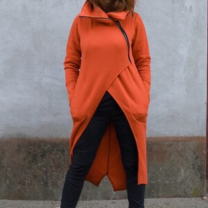 Op de afbeelding: Een lange, oranje hoodie met een rits aan de voorkant en twee grote zakken. De hoodie is in een wikkel-stijl en heeft een hoge kraag.