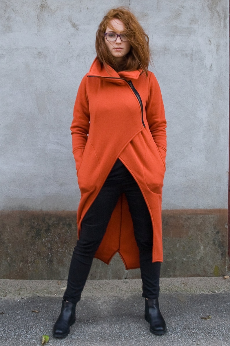 Op de afbeelding: Een lange, oranje hoodie met een rits aan de voorkant en twee grote zakken. De hoodie is in een wikkel-stijl en heeft een hoge kraag.