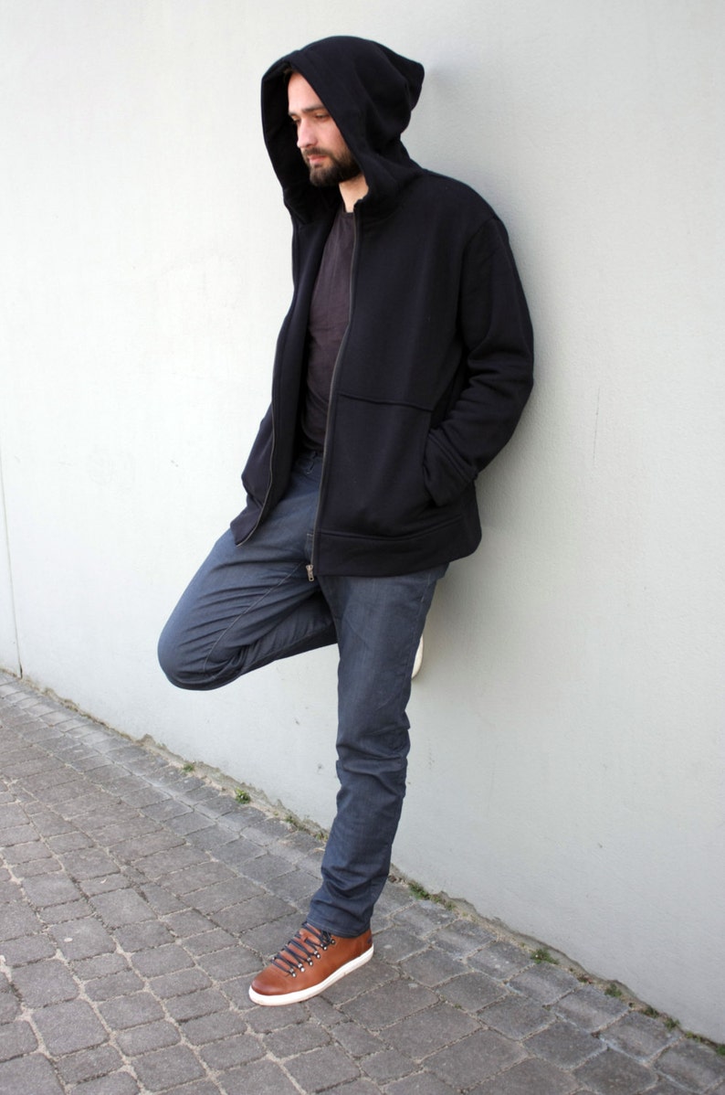 Pu&ograve; includere: Una felpa con cappuccio nera con chiusura a zip. Il modello indossa la felpa con il cappuccio su e sta in piedi con una gamba piegata. Il modello indossa jeans blu e scarpe di cuoio marroni.