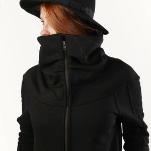 May include: A black zip-up jacket with a high, folded collar and long sleeves. The jacket has a zipper closure and a black fedora hat is worn on top of the head.