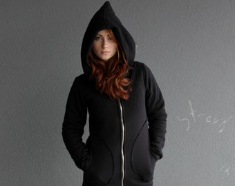 以下が含まれることがあります： ジッパー付きの黒いフード付きスウェットシャツを着た女性。フードをかぶっており、カメラを見ている。