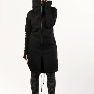 May include: A black long-sleeved jacket with a high collar and a zipper closure. The jacket has a relaxed fit and a slightly longer length. The model is wearing the jacket with black leggings and black boots.
