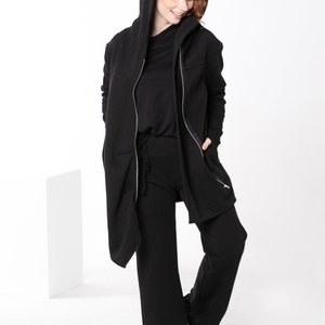May include: A black hooded jacket with a zipper closure and two zippered pockets. The jacket is worn over a black long-sleeved shirt and black pants. The person is wearing yellow sneakers.