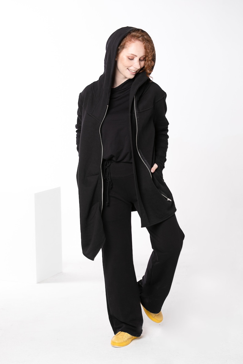 May include: A black hooded jacket with a zipper closure and two zippered pockets. The jacket is worn over a black long-sleeved shirt and black pants. The person is wearing yellow sneakers.