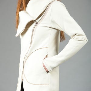 May include: A white zippered jacket with a large, folded collar. The jacket has a unique design with a curved seam on the front and a pocket on the left side.