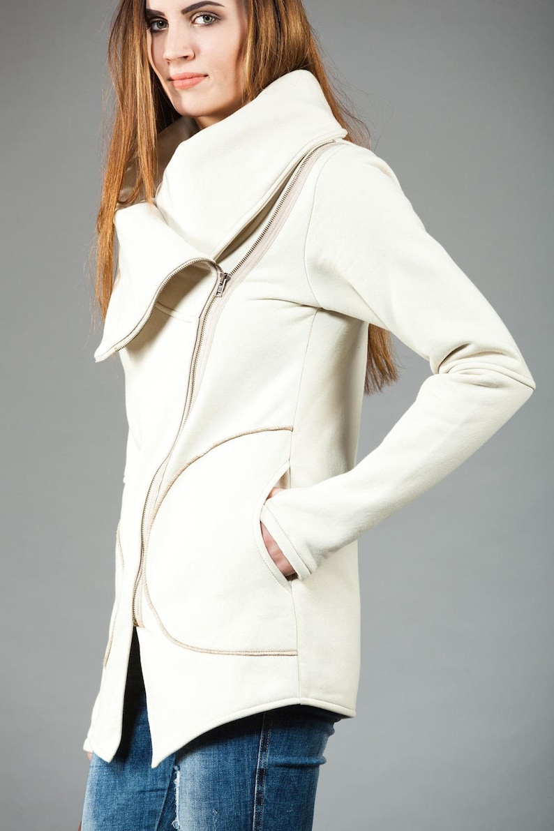 May include: A white zippered jacket with a large, folded collar. The jacket has a unique design with a curved seam on the front and a pocket on the left side.