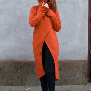 Op de afbeelding: Een lange, roestkleurige hoodie met een hoge hals en een lange split aan de voorkant. De hoodie heeft een zak aan de linkerkant en wordt gedragen met een zwarte broek en zwarte enkellaarsjes.