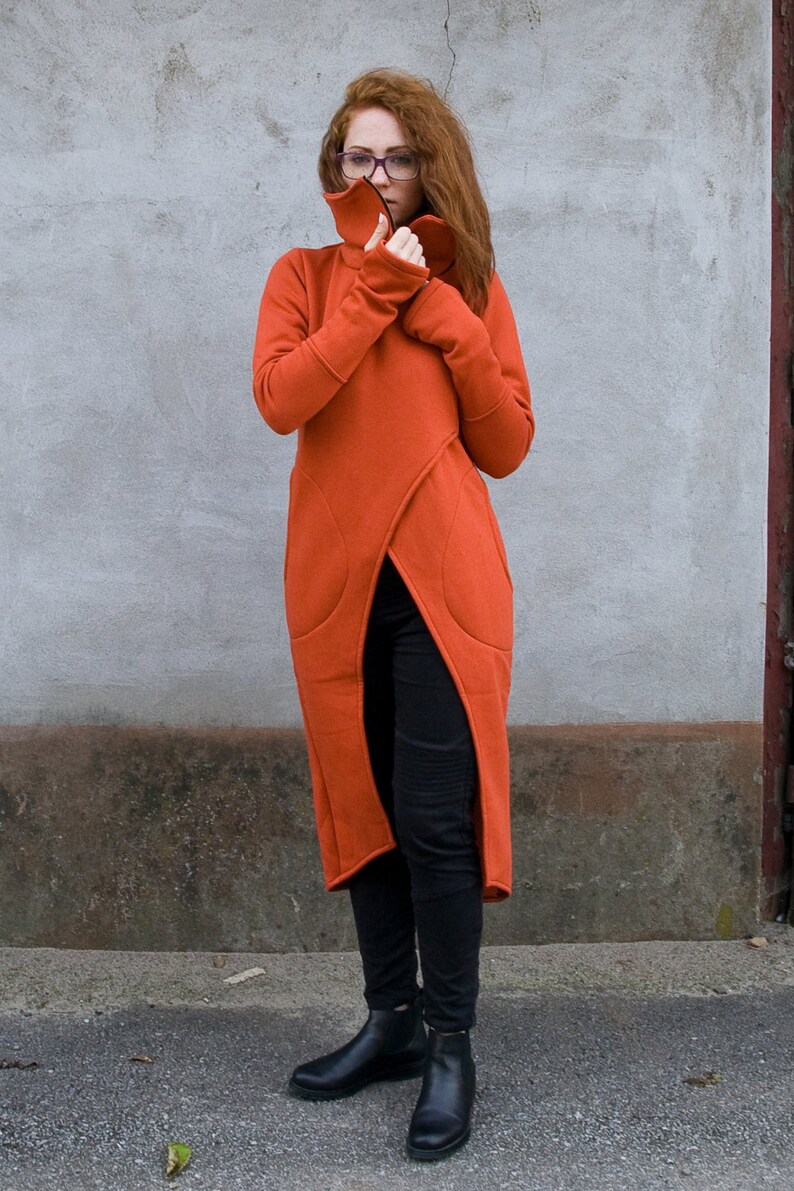 Op de afbeelding: Een lange, roestkleurige hoodie met een hoge hals en een lange split aan de voorkant. De hoodie heeft een zak aan de linkerkant en wordt gedragen met een zwarte broek en zwarte enkellaarsjes.