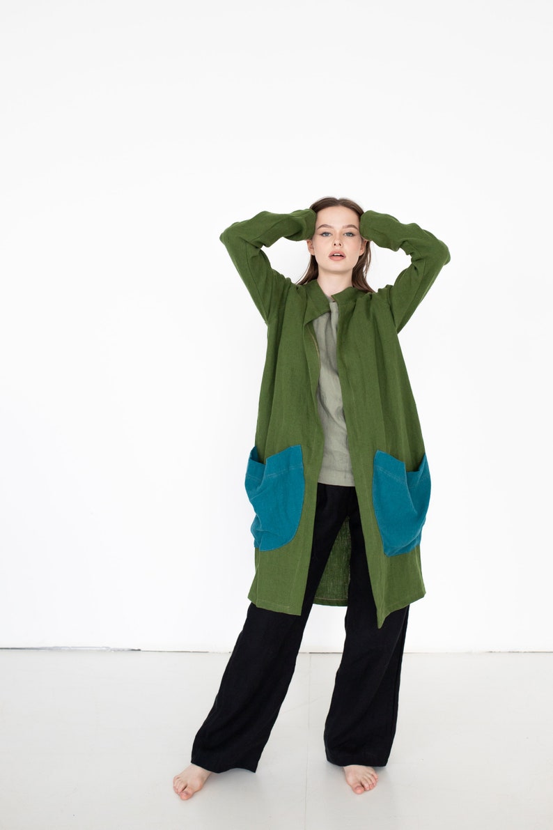 May include: A long, green linen coat with two large blue pockets. The coat is open and the person wearing it is standing with their hands on their head.