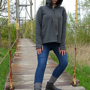 Pode incluir: Uma mulher vestindo um moletom cinza com capuz e z&iacute;per, jeans azul e botas pretas de tornozelo. Ela est&aacute; em p&eacute; em uma ponte de madeira.
