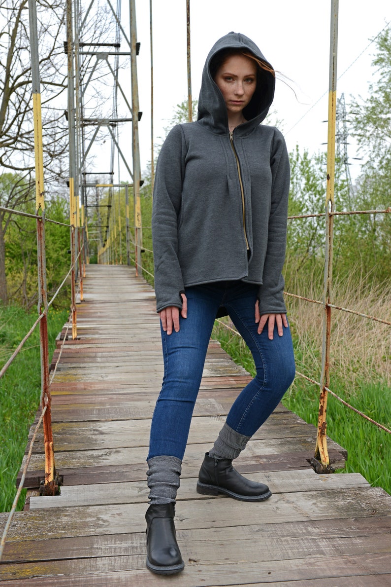 Pode incluir: Uma mulher vestindo um moletom cinza com capuz e z&iacute;per, jeans azul e botas pretas de tornozelo. Ela est&aacute; em p&eacute; em uma ponte de madeira.