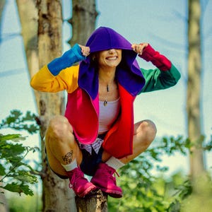 Könnte beinhalten: Bunte Kapuzenjacke mit lila Kapuze und roten, gelben, blauen und grünen Ärmeln. Die Person trägt ein weißes Oberteil, Shorts und rosa Stiefel. Die Person sitzt auf einem Baumstumpf im Wald.