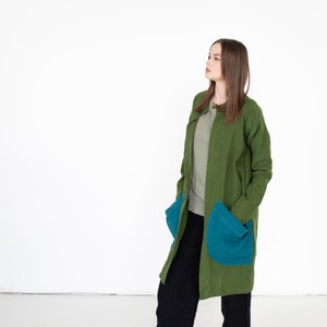May include: A woman wearing a long, green coat with large blue pockets. She is wearing black pants and is standing on a white background.