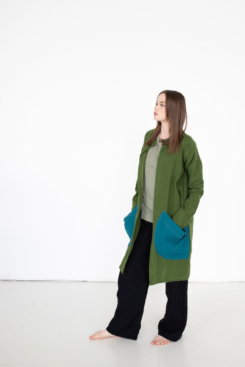 May include: A woman wearing a long, green coat with large blue pockets. She is wearing black pants and is standing on a white background.