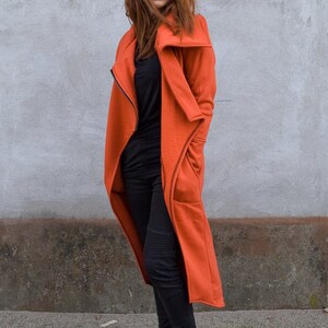 Op de afbeelding: Een vrouw draagt een lange, oranje jas met een rits en zakken. Ze draagt een zwarte broek en zwarte enkellaarsjes. Ze heeft rood haar en draagt een bril.