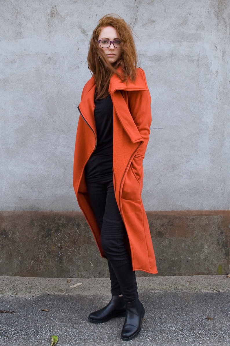 Op de afbeelding: Een vrouw draagt een lange, oranje jas met een rits en zakken. Ze draagt een zwarte broek en zwarte enkellaarsjes. Ze heeft rood haar en draagt een bril.