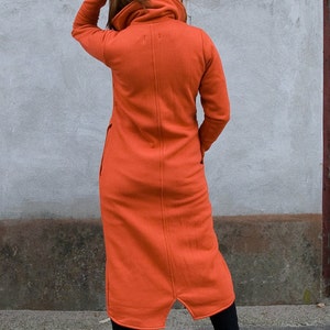 Op de afbeelding: Een lange, oranje, gekapte jurk met een hoge kraag en een lange, zwierige rok. De jurk is gemaakt van een zachte, comfortabele stof en heeft een ontspannen pasvorm.