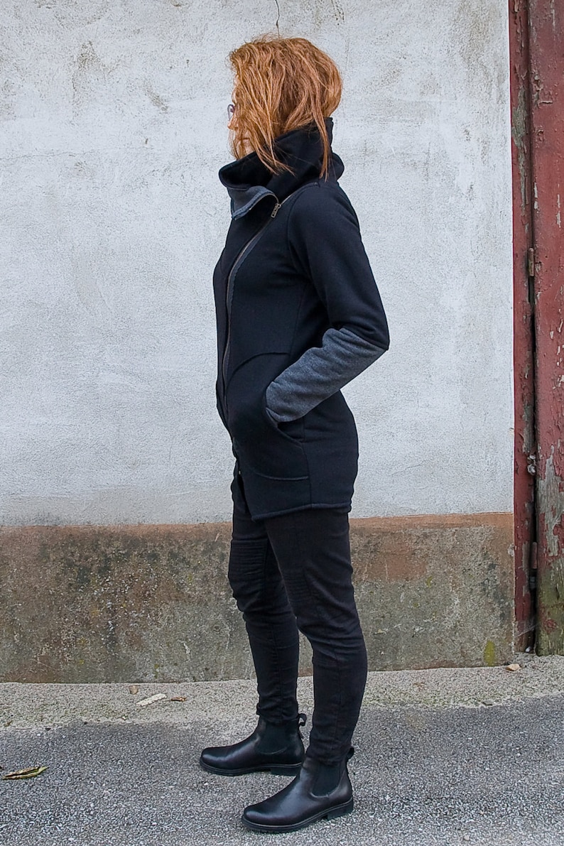 Pu&ograve; includere: Una giacca nera a maniche lunghe con cerniera e colletto alto. La giacca ha un pannello grigio sulla manica. La persona indossa pantaloni neri e stivaletti neri.
