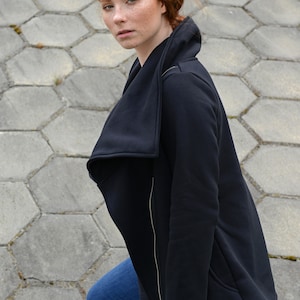 May include: A woman with red hair wearing a black zip-up jacket with a large, folded collar. The jacket has a zipper down the front and a pocket on the left side. She is wearing blue jeans.