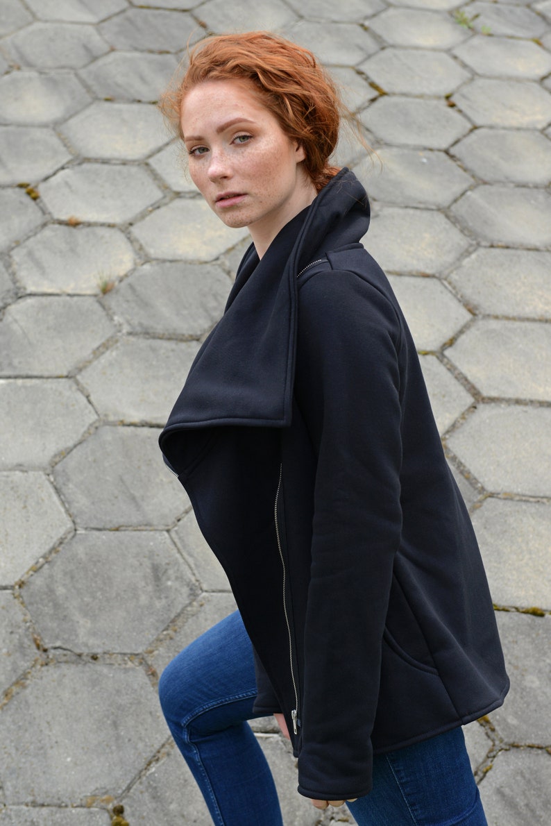 May include: A woman with red hair wearing a black zip-up jacket with a large, folded collar. The jacket has a zipper down the front and a pocket on the left side. She is wearing blue jeans.